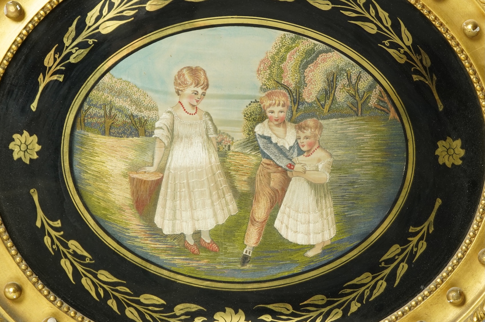 A large oval early 20th century embroidery of three children in a landscape. Worked in mostly stem stitch with knotting embroidery on the trees. The embroidery framed in Verre Eglomise glass and a wide wooden gilt frame